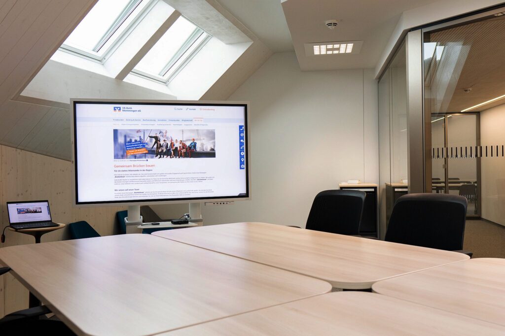 Smartboard & Medientechnik für kreative ZusammenarbeitDieser Huddle Room kombiniert Smartboard-Funktion mit kabelloser Präsentation. Die integrierte Medientechnik unterstützt agile Workshops, spontane Ideenfindung und interaktive Gruppenarbeit – perfekt für moderne Besprechungsräume. Glas-Meetingraum der Volksbank Memmingen eG mit einem Tisch und zwei Stühlen