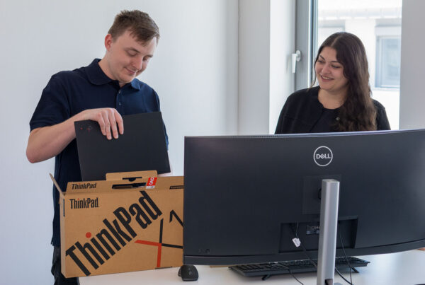 Zwei Personen die hinter einem Monitor stehen und einer einen Laptop auspackt.